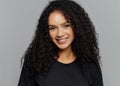 Optimistic curly young woman with gentle smile, looks positively at camera, wears casual black clothes, isolated on grey