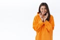 Optimistic good-looking modern girl in orange hoodie peeking at camera while cross fingers good luck and making wish