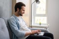 Optimistic man sitting on the couch and using laptop computer for messaging. Happy businessman working remotely in Royalty Free Stock Photo