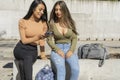 Two young women listening to music with their cell phone sitting on a concrete bench in a plaza. Technology, communications, joy Royalty Free Stock Photo