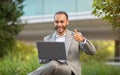 Optimistic young businessman in suit working on laptop outdoors, showing thumb up Royalty Free Stock Photo