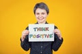 Optimistic young woman hipster holding a white paper sheet with the message text think positive isolated over yellow background.