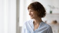 Optimistic young woman standing indoors looking at window smiling dreaming