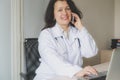 Optomistic doctor woman in medical institution sits at the desktop with computer and speaks on the phone. Phisician is smiling