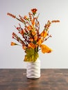 Orange artificial flowers in a white vase sitting on a wooden cutting board with a white wall behind. Fall home decor for autumn