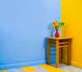 Orange azalea rhododendron flower in a tiny vase on a stool in a corner in super bright and contrast room with yellow blue painted