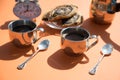 On an orange background on a Sunny morning, there are metal coffee cups, a coffee pot, buns on a plate, and an alarm clock.