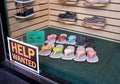 An orange and black help wanted sign in a storefront window with background display of summer shoes.