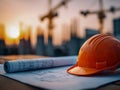 Orange construction helmet resting on architectural blueprints with cranes and building structures silhouetted against a warm Royalty Free Stock Photo