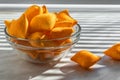 Orange corn chips in the form of pillows lie in a glass bowl on a white background, consecrated by the sun`s rays passing through