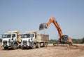 orange crawler excavator and two gray construction dump trucks in the process of loading and transporting soil