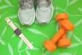 Orange dumbbells, women`s sneakers and a fitness watch to control your health on a green background