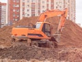 An orange excavator on a construction site against the background of a multi-storey building digs a trench. Royalty Free Stock Photo