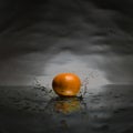 An orange falling and making a splash in liquid water on a reflective mirrored floor with a moody background.