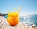 Orange fruit as the cup with orange juice splash and straw on sandy beach. Blue sparkling sea at the background. Drink concept Royalty Free Stock Photo