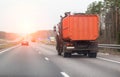 An orange garbage truck drives along an asphalt country road in the spring against the backdrop of sunset. Garbage