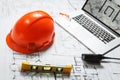 Orange hard hat, laptop with drawings, glasses and walkie talkie with blueprints on a table