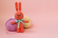 Orange knitted hare stands on the background of multi-colored balls of yarn on a pink background. There is a place for text