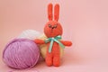 Orange knitted hare stands on the background of multi-colored balls of yarn on a pink background. There is a place for text