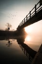 Orange misty foggy sunrise from from under wooden bridge over river reflected in canal. Beautiful reflection in still tranquil. Royalty Free Stock Photo