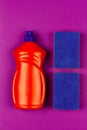 Orange plastic bottle of dishwashing liquid and blue sponge on purple background close-up