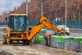 An orange road excavator unloads stacks of paving slabs to the side of a road along a city street on a spring afternoon