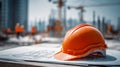 Orange safety helmet resting on architectural blueprints at an active urban construction site with blurred workers and cranes in Royalty Free Stock Photo