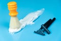 An orange shaving brush stands in whipping foam and two black razors on a blue background