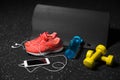 Orange sport shoes, yellow dumb-bells, pilates mat, blue bottle, and phone with headphones on a black background. Sport