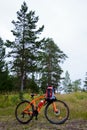 An orange sports bike with hiking backpack stands in a pine forest, on a cloudy day. Vertical Travel Photo