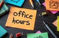 Orange sticky note with bold black text OFFICE HOURS on black background. Colorful sticky notes pens and clock scattered Royalty Free Stock Photo
