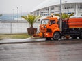 An orange watering machine washes the road on a cloudy day against the background of the stadium and palm trees. Men at