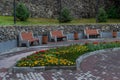 orange wooden bench with trash cans nearby, on stone path with flower beds with yellow and red flowers in park alleys