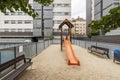 An orange wooden and synthetic material slide on a sand surface and wooden and metal benches in a communal area of residential