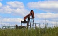 Orange working pump jack on oil or gas well on horizon of green field with jug of chemicals sitting beside it and grass and wild f Royalty Free Stock Photo