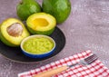 Organic avocado with seed, avocado halves, and fresh guacamole in a bowl on a black plate with cloth and cutlery on a gray stone Royalty Free Stock Photo