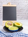 Organic avocado with seed, avocado halves, and fresh guacamole in a bowl on black plate with a small blackboard and cloth on Royalty Free Stock Photo
