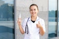 Oriental smiling female nurse with stethoscope in medical gown holding syringe up and pointing thumbs up Royalty Free Stock Photo