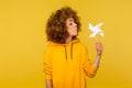 Origami hand mill. Portrait of carefree childish curly-haired woman in hoodie blowing at paper windmill