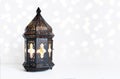 Ornamental dark Moroccan, Arabic lantern on the white table. Burning candle, glittering bokeh lights stars. Greeting