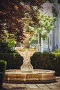 Ornate water fountain without water sits on pavement with hedges and japanese maple and blurred cottage in background