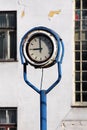 Outdoor analog clock with rusted metal frame in middle of abandoned industrial complex with old dilapidated building in background