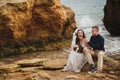 Outdoor beach wedding ceremony near the sea, stylish happy smiling groom and bride are sitting on stones, having fun and Royalty Free Stock Photo