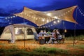Outdoor camping tent in the forest park, party dinner with friends under tarp or flysheet and warm lighting at night near natural