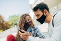 Outdoor candid portrait of a young man and a woman sitting outside looking to the smartphone. Two young students in protective Royalty Free Stock Photo