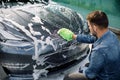 Outdoor car wash self service. Back angle view of handsome young Caucasian man washing luxury gray electric car with Royalty Free Stock Photo