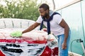 Outdoor car wash self service. Handsome young African man washing redcar with green brush and foam Royalty Free Stock Photo