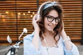 Outdoor close-up portrait of cute smiling girl in glasses touching white earphones. Fascinating young woman with dark Royalty Free Stock Photo