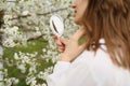 Outdoor close up portrait of young beautiful woman in blooming garden on spring day. Model looking in little mirror, posing in Royalty Free Stock Photo