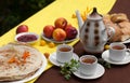 An outdoor composition with tea cups, a tea pot, a plate of pancakes, pastry, ripe fruit and field flowers on a bright table cloth Royalty Free Stock Photo